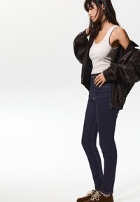 Temno modri visoki pasovi jeans s kontrastnim šivanjem, kombinirani z oprijetim belim topom in velikim rjavim usnjenim jopičem.