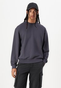 Grå sweatshirt med långa ärmar och rundad hals, med enfärgad design och ribbad nederkant. Matchas med svarta cargobyxor och en svart keps.