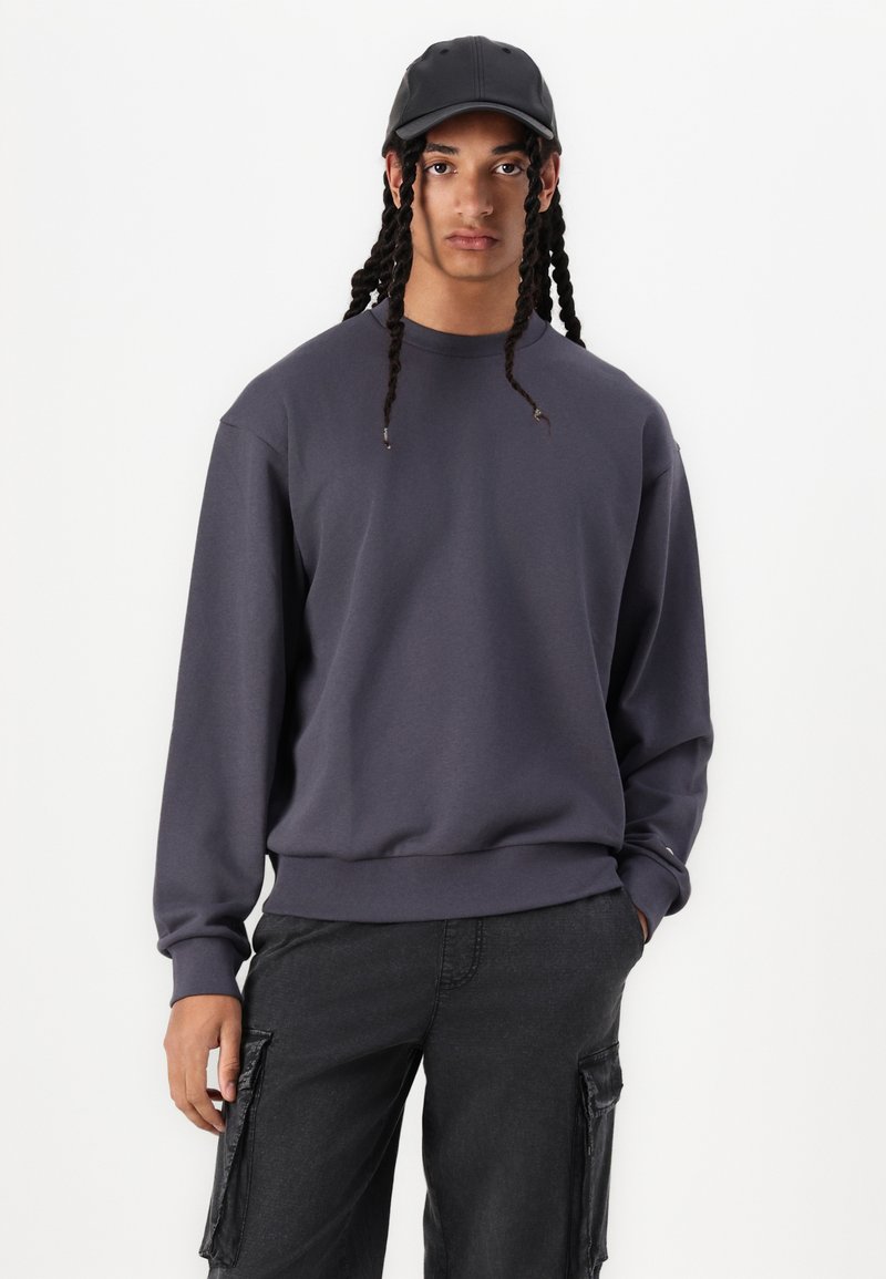 Grå sweatshirt med långa ärmar och rundad hals, med enfärgad design och ribbad nederkant. Matchas med svarta cargobyxor och en svart keps.