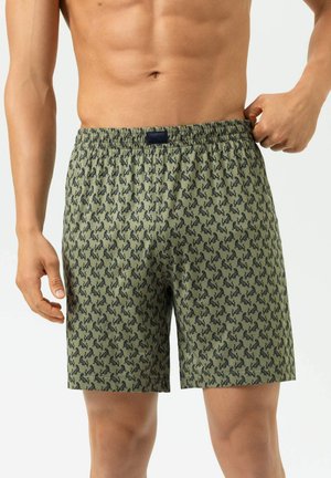 Mann trägt olivgrüne Boxershorts mit schwarzem Katzenmuster, zieht den Bund leicht nach außen und zeigt Vorderkörper und obere Beine.