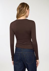 Top marrone a maniche lunghe con design aderente, texture liscia e scollo rotondo, abbinato a jeans in denim blu scuro con tasche posteriori.