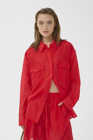 Junge Frau mit hellbraunen Haaren, die ein lockeres, knallrotes Hemd mit Knöpfen und passende Shorts trägt, steht vor einem weißen Hintergrund.