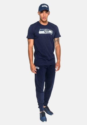 Man wearing navy blue Seattle Seahawks cap, t-shirt with logo, matching joggers, and dark sneakers, standing against plain background.