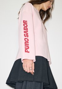 Rózsaszín hosszú ujjú ing, vörös "PURO SABOR" felirattal az ujján, sötét pliszírozott szoknyával és ezüst gyűrűkkel párosítva.