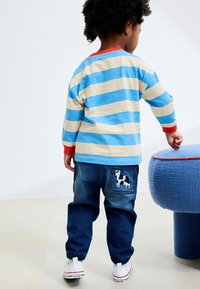 Langærmet skjorte med striber i blå og beige med røde accenter, kombineret med mørkeblå joggers med en ko-broderi på baglommen.