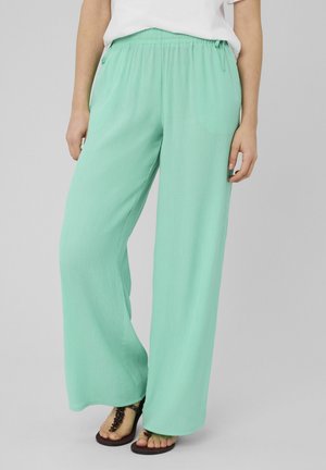 Femme portant un pantalon ample vert menthe à taille élastique et des sandales noires, debout devant un fond uni.