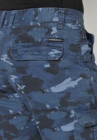 Zbliżenie na niebieskie i czarne spodenki cargo w kamuflażowym wzorze z klapkowymi kieszeniami i etykietą "KRSDenim" z tyłu.