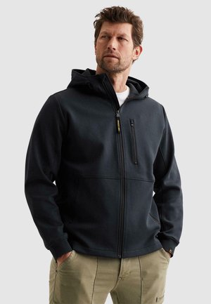 Mann trägt eine schwarze Kapuzenjacke mit Reißverschluss und Taschen sowie eine braune Hose, schaut zur Seite vor einem schlichten Hintergrund.