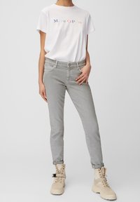 Bílá bavlněná tričko s vícebarevným textem "Marc O'Polo", v kombinaci se světle šedými slim-fit džíny a béžovými šněrovacími botami.