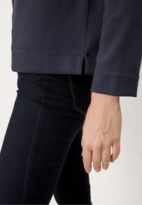 Camicia a maniche lunghe blu scuro con fenditure laterali, abbinata a jeans scuri caratterizzati da cuciture chiare lungo le cuciture. Focus sulle trame dei tessuti e sulla vestibilità.
