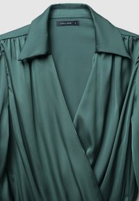 Blusa avvolgente in satin verde smeraldo con colletto, caratterizzata da dettagli plissettati sulle spalle e una texture liscia. L'etichetta riporta "CALLIOPE S".