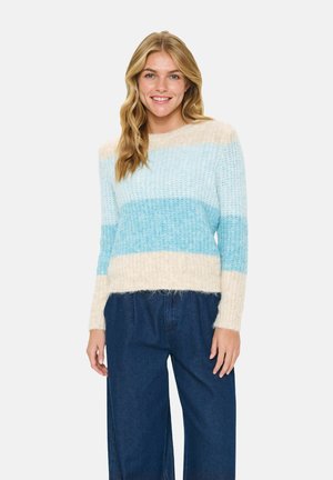 Smiling woman wearing a fuzzy knit sweater with cream, light blue, and turquoise horizontal stripes and dark blue wide-leg jeans.