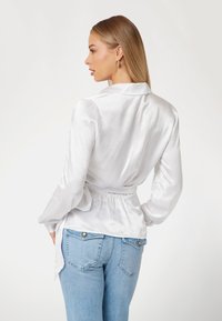 Balti atlasinė bluzė su ilgomis rankovėmis ir surišta juosmena, derinama su šviesiai mėlynomis džinsais, turinčiais dekoratyvinius kišenes gale.