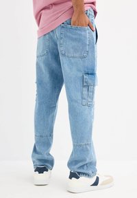 Pantalones cargo de denim azul claro con múltiples bolsillos, piernas ligeramente entalladas y costuras visibles. Combinados con zapatillas blancas con acentos en azul marino.