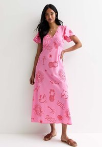 Rosa maxiklänning med korta volangärmar som har röda tryckta mönster av sol, vågor, ananaser och abstrakta mönster. Platta sandaler bärs.