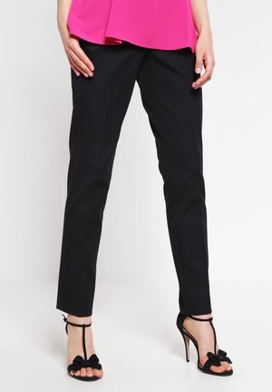 Frau trägt schwarze Slim-Fit-Hose, schwarze High-Heel-Sandalen mit Schleifen und ein knallpinkes Top und steht vor weißem Hintergrund.