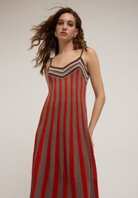Robe tricotée avec des rayures verticales rouges et brunes, bretelles spaghetti et un décolleté en chevron. Texture douce et coupe décontractée.