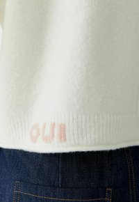 Weißer Strickpullover mit dezenten rosa Buchstaben nahe dem gerippten Saum, kombiniert mit dunkelblauen Jeans aus Denim mit sichtbaren Nähten und Gesäßtasche.