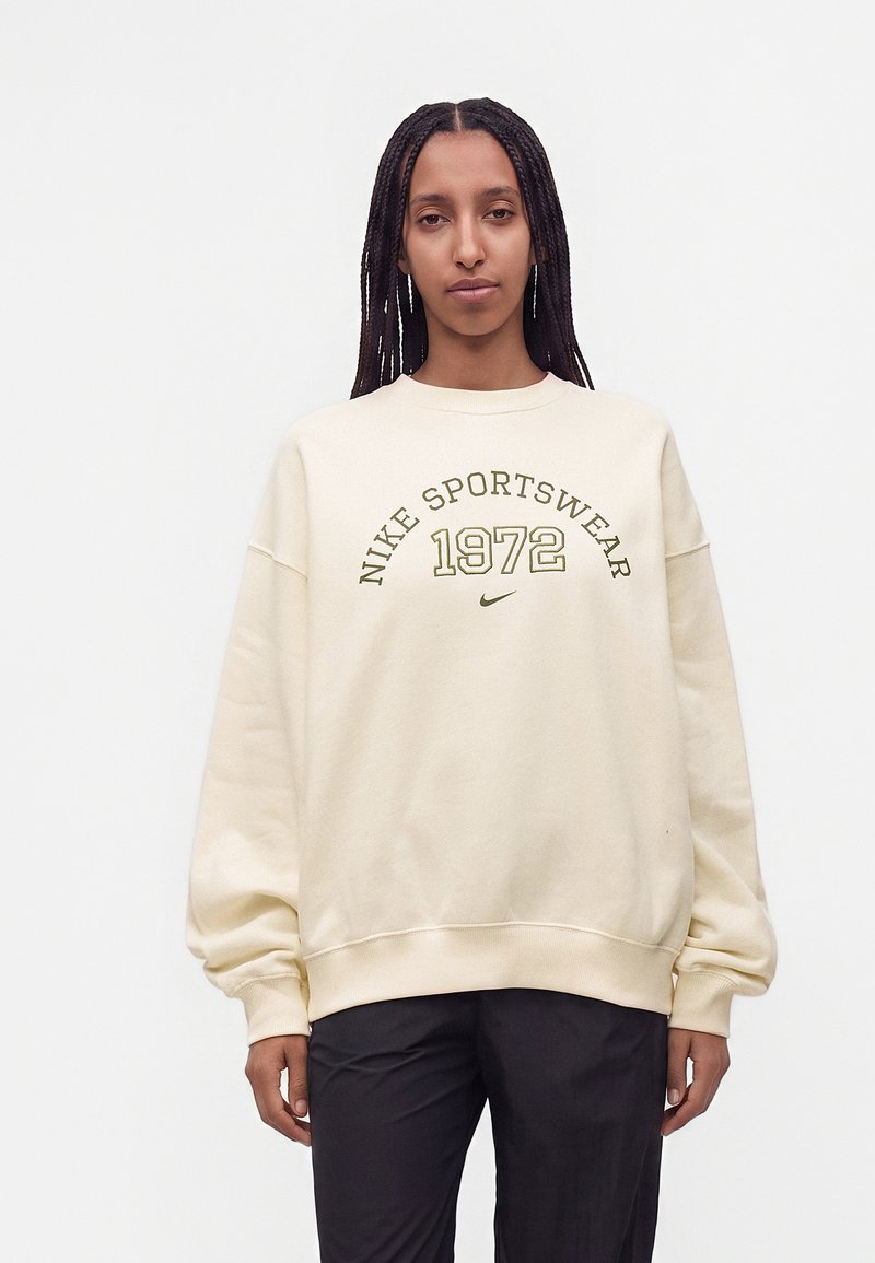 Femme aux longues tresses portant un sweat-shirt crème Nike Sportswear avec "1972" imprimé à l'avant et un pantalon noir.