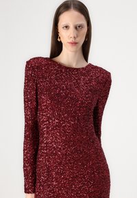 Red sequin dress with long sleeves and a rounded neckline. Features a fitted silhouette and textured surface from overlapping sequins.