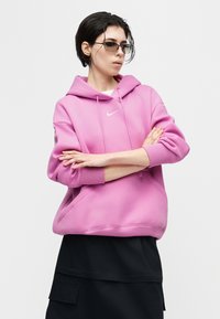 Άτομο με κοντά σκούρα μαλλιά που φοράει γυαλιά ηλίου και ροζ φούτερ Nike, με σταυρωμένα τα χέρια, στέκεται μπροστά σε απλό λευκό φόντο.