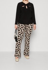 Top nero a maniche lunghe con dettagli cut-out, abbinato a pantaloni a zampa d'elefante con stampa leopardata. La modella tiene in mano una piccola borsa nera e indossa scarpe color crema.