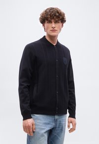 Νέος άνδρας με σγουρά μαλλιά, φορώντας μαύρο ζακέτα με κουμπιά και ανοιχτόχρωμο μπλε τζιν, στέκεται μπροστά σε απλό ανοιχτό φόντο.