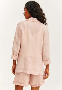 Světle růžový lněný dvoudílný komplet, který se skládá z košilové halenky s límcem a dlouhým rukávem a šortek. Rukávy jsou ohrnuté a látka má texturovaný povrch.