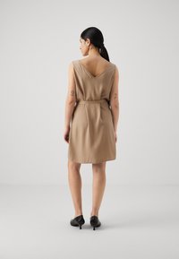 Beige uten ermer kjole med dyp V-hals bak, innsvingt midje og flytende stoff. Båret med svarte hæler, mot en enkel grå bakgrunn.