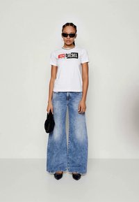 T-shirt blanc à manches courtes avec un logo imprimé noir et rouge, associé à un jean en denim bleu clair à jambes larges. Les accessoires comprennent des lunettes de soleil noires et un petit sac noir.