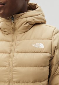 Chaqueta acolchada de color marrón claro con un alto cuello, cierre de cremallera y logo en blanco. Presenta una textura de costura horizontal y una tela suave.