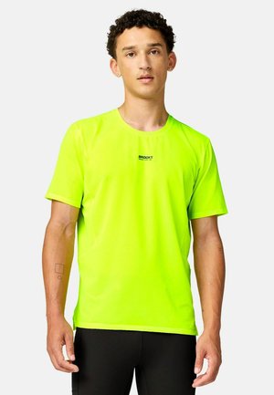Giovane uomo con capelli ricci che indossa una maglietta Brooks giallo neon brillante e pantaloni neri, in piedi su uno sfondo chiaro uniforme.