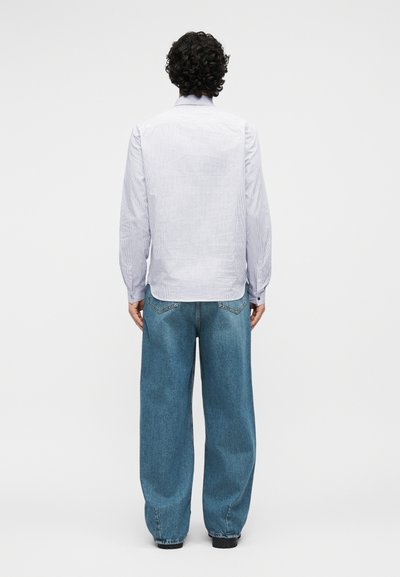 Άνδρας με σγουρά μαλλιά που φοράει ανοιχτό μπλε πουκάμισο με μακρύ μανίκι και φαρδιά μπλε τζιν, στέκεται μπροστά σε λευκό φόντο με τα χέρια χαλαρά.