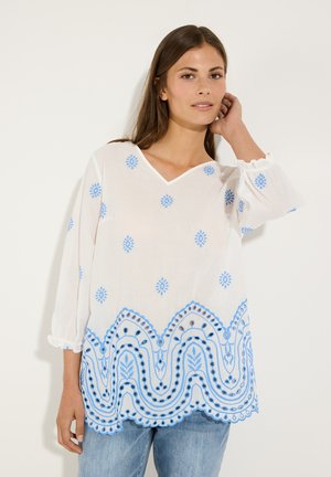 Femme aux longs cheveux foncés portant un chemisier blanc avec une broderie florale bleue et un bord à pompons, associé à un jean bleu clair.