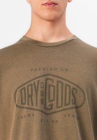 Nærbilde av en person som har på seg en brun T-skjorte med teksten "Premium Co. Dry Goods Jack & Jones Est. 90" trykt på forsiden.