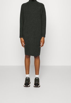 Person iført en mørkegrå, langærmet, knælængde sweaterkjole med hvide sokker og sorte sneakers stående mod en ensfarvet hvid baggrund.