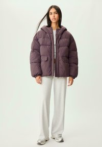 Dunkelviolette Pufferjacke mit Kapuze, zwei vorderen Taschen und glatter Textur, kombiniert mit hellgrauen Sweatpants und metallischen Sneakers.