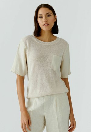 Frau mit schulterlangen dunklen Haaren, die einen beigen Stricktop mit Tasche und passende hellfarbige Hose trägt, steht vor einem einfarbigen Hintergrund.