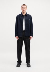 Camisa azul-marinho abotoada com um bolso no peito, combinada com calças de ganga escuras e sapatos pretos. Ajuste casual, textura suave, design simples.