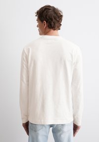 Langärmliges Hemd in hellem Beige, aus weichem Stoff hergestellt. Verfügt über einen Rundhalsausschnitt, geraden Schnitt und minimale Nähdetails auf der Rückseite.