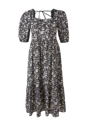 Kvinde iført en sort blomstret midikjole med pufærmer og beige chunky sneakers mod en ensfarvet lys baggrund.