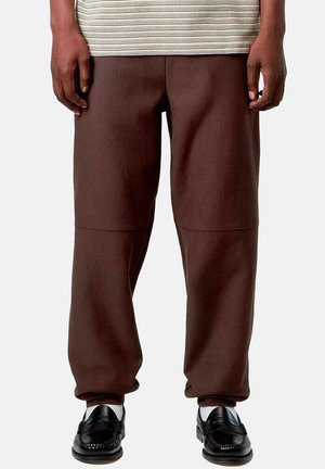Hombre que lleva pantalones marrones de corte holgado, calcetines blancos y zapatos negros sin cordones, de pie contra un fondo liso y claro.
