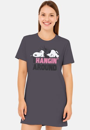 Junge Frau lächelnd, trägt ein dunkelgraues Kleid mit entspannter liegender Snoopy-Grafik und dem Text "Just Hangin' Around" in Pink und Schwarz.