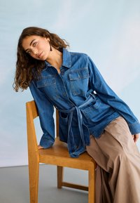 Femeie cu păr creț purtând o jachetă din denim albastru și pantaloni largi bej, stând pe scaun din lemn, cu corpul orientat lateral, pe un fundal deschis.