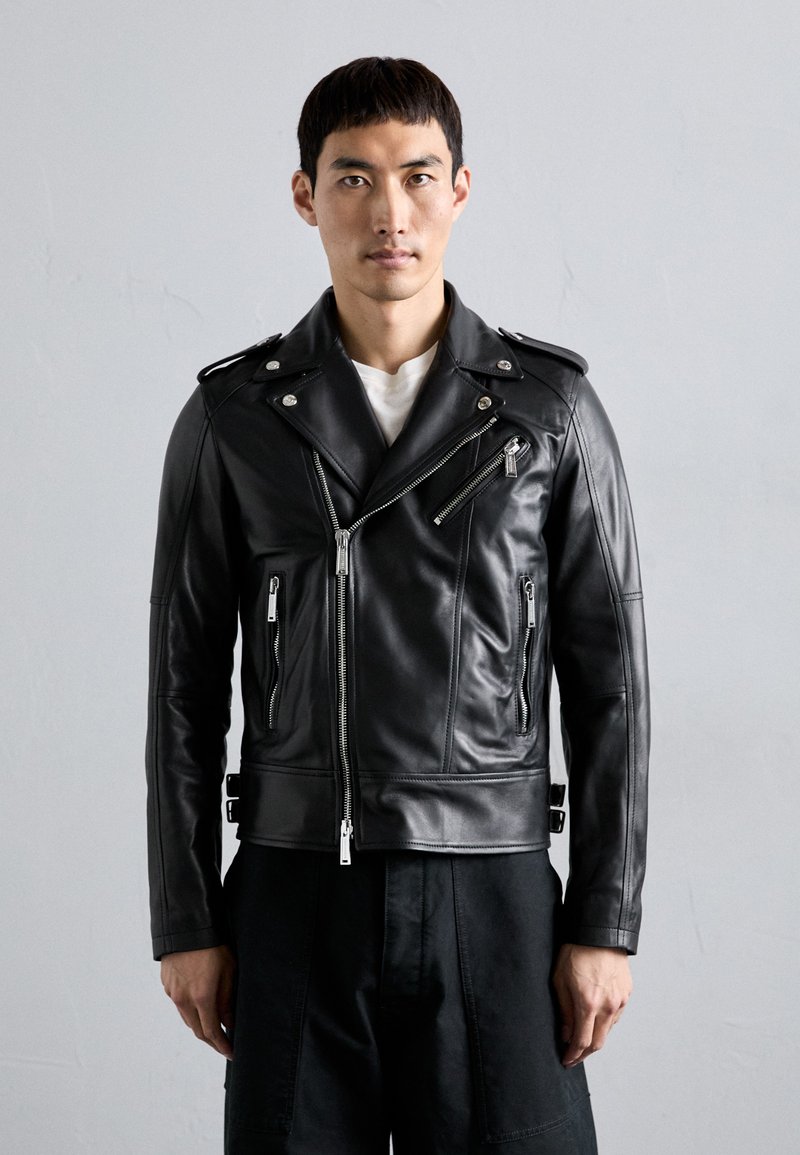 Giacca da biker in pelle nera con cerniere e accessori in argento, dotata di una cerniera frontale inclinata, tasche laterali e un design aderente.