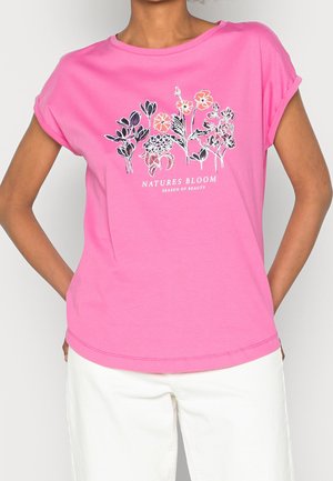 Osoba ubrana w różowy t-shirt z czarno-pomarańczowymi kwiatowymi wzorami oraz białym napisem "Natures Bloom Season of Beauty", zestawiona z białymi spodniami.