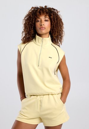 Vrouw met krullend haar draagt een lichtgeel mouwloos topje met rits en bijpassende shorts, staand met handen in de zakken tegen een effen achtergrond.