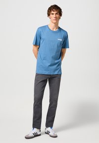 Camiseta de algodón azul con cuello redondo, que presenta un pequeño logo blanco de "Wrangler" en el pecho, combinada con jeans grises y zapatillas multicolores.