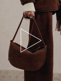 Hnědá kabelka z broušené kůže s zakřiveným designem a hnědými koženými popruhy, která má hladkou texturu a polostrukturovaný tvar.