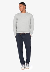 Sudadera gris texturizada con puños acanalados, pantalones azul marino y zapatillas ligeras. Presenta un logo en el pecho y un cinturón estampado con reloj.